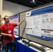 A man in a red polo shirt with a conference lanyard is presenting a research poster to a woman in a U.S. Army military uniform with Airborne insignia at what appears to be a scientific or military research conference. The poster is titled 'SeptiBurnAlert: AI/ML tool integrated with qPCR assay to score the risk of burn sepsis from the first blood draw after ICU admission' and is affiliated with DHA (Defense Health Agency) and WRAIR. A third person is partially visible in the background.