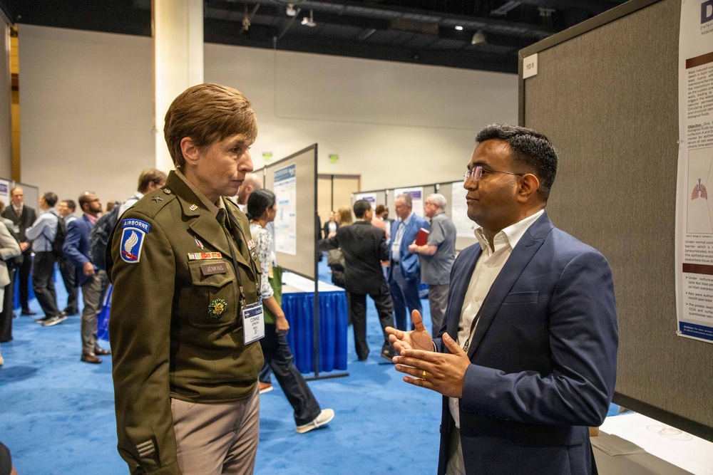 Two individuals are engaged in conversation at what appears to be a professional or academic conference with research poster displays. One person is wearing a U.S. Army uniform with an Airborne patch and visible rank insignia, while the other is dressed in a navy blue blazer. The setting is a large convention hall with blue carpet and multiple poster presentation boards visible in the background.