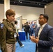 Two individuals are engaged in conversation at what appears to be a professional or academic conference with research poster displays. One person is wearing a U.S. Army uniform with an Airborne patch and visible rank insignia, while the other is dressed in a navy blue blazer. The setting is a large convention hall with blue carpet and multiple poster presentation boards visible in the background.