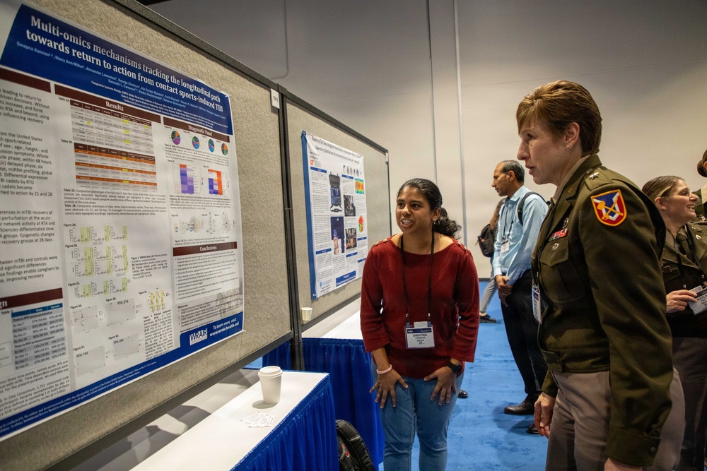 A scientific poster session at what appears to be a military or defense-related research conference. A civilian researcher in a red sweater is presenting her research poster on 'Multi-omics mechanisms tracking the longitudinal path towards return to action from contact sports-induced TBI' to a military officer in Army dress uniform. The setting is a conference hall with blue carpeting and multiple poster displays.
