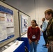 A scientific poster session at what appears to be a military or defense-related research conference. A civilian researcher in a red sweater is presenting her research poster on 'Multi-omics mechanisms tracking the longitudinal path towards return to action from contact sports-induced TBI' to a military officer in Army dress uniform. The setting is a conference hall with blue carpeting and multiple poster displays.