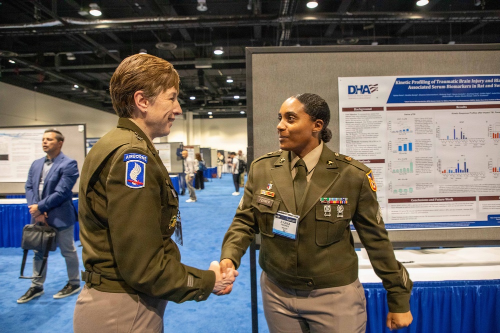 Two U.S. Army soldiers in Army Green Service Uniforms are shaking hands at what appears to be a military medical or scientific conference. A research poster behind them displays a DHA (Defense Health Agency) study titled 'Kinetic Profiling of Traumatic Brain Injury and Blast-Associated Serum Biomarkers in Rat and Swine.' The setting is a large convention hall with multiple poster displays and other attendees visible in the background.