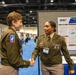 Two U.S. Army soldiers in Army Green Service Uniforms are shaking hands at what appears to be a military medical or scientific conference. A research poster behind them displays a DHA (Defense Health Agency) study titled 'Kinetic Profiling of Traumatic Brain Injury and Blast-Associated Serum Biomarkers in Rat and Swine.' The setting is a large convention hall with multiple poster displays and other attendees visible in the background.