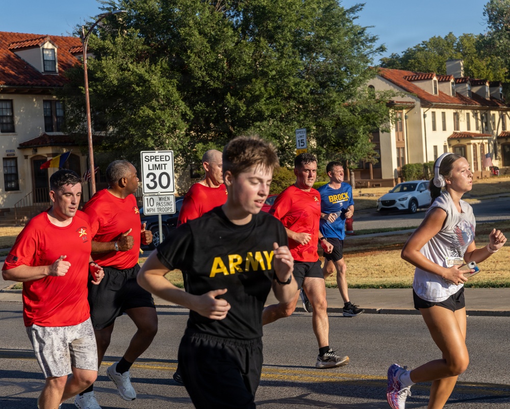 A group of runners are jogging along a road on what appears to be a military installation, based on the uniform-style buildings with red tile roofs and the speed limit sign reading '10 MPH When Passing Troops.' Several participants wear red shirts with a military insignia, one wears a black 'ARMY' shirt, and a civilian female runner with headphones runs alongside them. The setting appears to be a morning run in warm, sunny conditions.