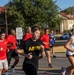 A group of runners are jogging along a road on what appears to be a military installation, based on the uniform-style buildings with red tile roofs and the speed limit sign reading '10 MPH When Passing Troops.' Several participants wear red shirts with a military insignia, one wears a black 'ARMY' shirt, and a civilian female runner with headphones runs alongside them. The setting appears to be a morning run in warm, sunny conditions.