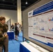 Two U.S. Army soldiers in military uniforms are viewing a scientific research poster at what appears to be a military medical or defense health conference. The poster, displayed on a board, presents research on 'Kinetic Profiling of Traumatic Brain Injury and Blast Overpressure Associated Serum Biomarkers in Rat and Swine Models' under the DHA (Defense Health Agency) banner. The setting is a large convention hall with other attendees and poster displays visible in the background.