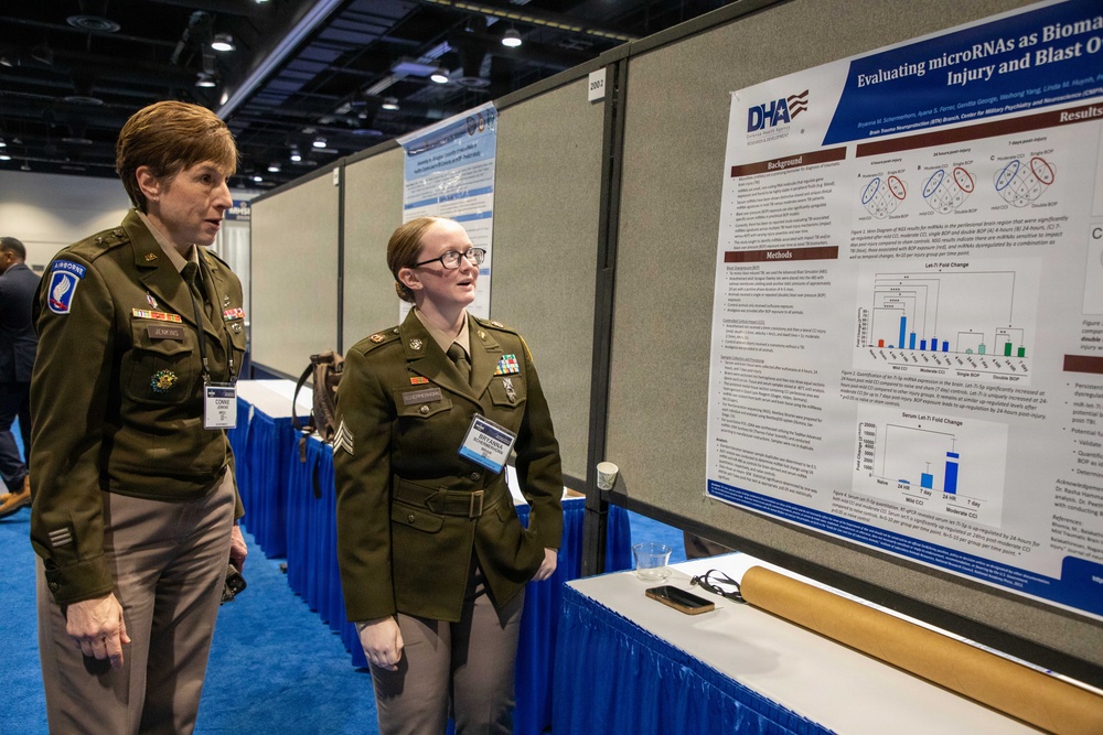 Two U.S. Army personnel in olive green dress uniforms are viewing a scientific research poster at what appears to be a military medical or neuroscience conference. The poster, affiliated with the Defense Health Agency (DHA), is titled 'Evaluating microRNAs as Bioma[rkers]' related to brain injury and blast exposure. The setting is a professional conference hall with blue-skirted display tables.