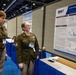 Two U.S. Army personnel in olive green dress uniforms are viewing a scientific research poster at what appears to be a military medical or neuroscience conference. The poster, affiliated with the Defense Health Agency (DHA), is titled 'Evaluating microRNAs as Bioma[rkers]' related to brain injury and blast exposure. The setting is a professional conference hall with blue-skirted display tables.
