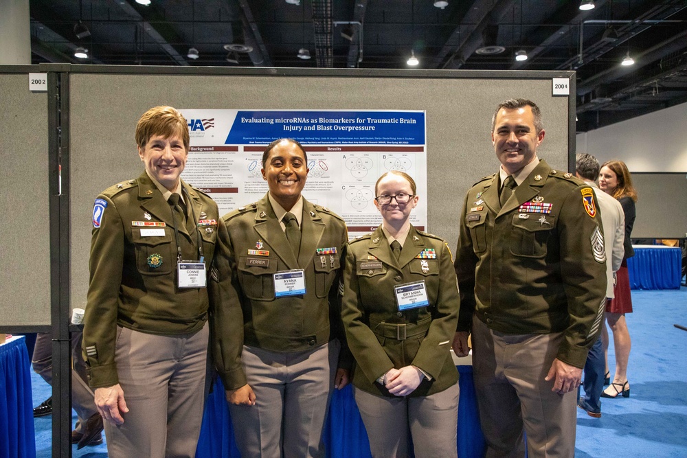 Four U.S. Army personnel in Army Service Uniforms (ASUs) pose together at what appears to be a military medical or scientific conference. They are standing in front of a research poster titled 'Evaluating microRNAs as Biomarkers for Traumatic Brain Injury and Blast Overpressure.' Name badges are visible on three individuals: Connie, Ayana Ferrer, and Bryanna.