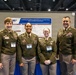 Four U.S. Army personnel in Army Service Uniforms (ASUs) pose together at what appears to be a military medical or scientific conference. They are standing in front of a research poster titled 'Evaluating microRNAs as Biomarkers for Traumatic Brain Injury and Blast Overpressure.' Name badges are visible on three individuals: Connie, Ayana Ferrer, and Bryanna.