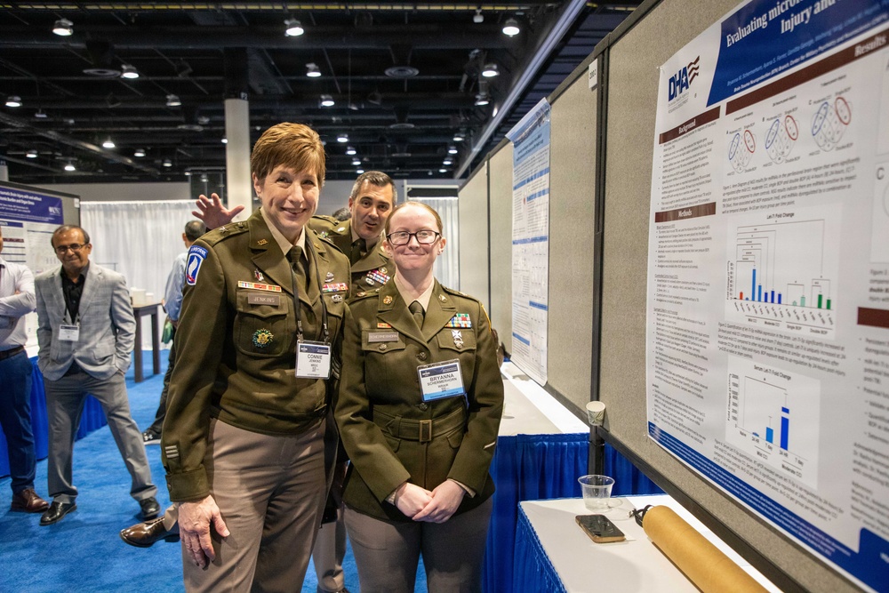 Two U.S. Army personnel in olive dress uniforms pose in front of a DHA (Defense Health Agency) research poster at what appears to be a military medical or scientific conference. A third uniformed individual is visible in the background. The poster is titled 'Evaluating micro... Injury and...' and contains charts, graphs, and research data.