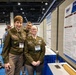 Two U.S. Army personnel in olive dress uniforms pose in front of a DHA (Defense Health Agency) research poster at what appears to be a military medical or scientific conference. A third uniformed individual is visible in the background. The poster is titled 'Evaluating micro... Injury and...' and contains charts, graphs, and research data.