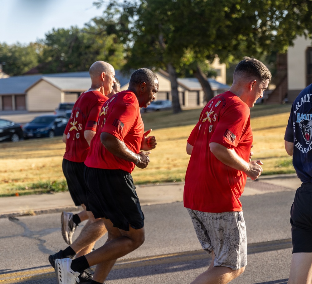 A group of individuals are running together on a road, with most wearing matching red t-shirts featuring military-style insignia and patches on the back. The setting appears to be a military installation or base, evidenced by the uniform shirts, visible 'ARMY' text on one participant's shirt, and the institutional buildings in the background. The run appears to be an organized group physical training or unit run event.