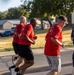 A group of individuals are running together on a road, with most wearing matching red t-shirts featuring military-style insignia and patches on the back. The setting appears to be a military installation or base, evidenced by the uniform shirts, visible 'ARMY' text on one participant's shirt, and the institutional buildings in the background. The run appears to be an organized group physical training or unit run event.