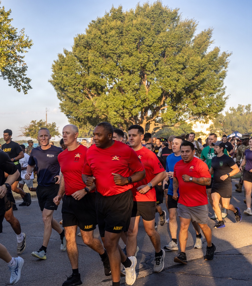A large group of people participating in an organized run or fun run event, likely on a military installation. Several participants in the foreground are wearing matching red shirts with a gold star insignia and black shorts labeled 'ARMY', suggesting U.S. Army affiliation. The event appears to take place on a paved road lined with mature trees during early morning or late afternoon light.