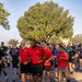 A large group of people participating in an organized run or fun run event, likely on a military installation. Several participants in the foreground are wearing matching red shirts with a gold star insignia and black shorts labeled 'ARMY', suggesting U.S. Army affiliation. The event appears to take place on a paved road lined with mature trees during early morning or late afternoon light.