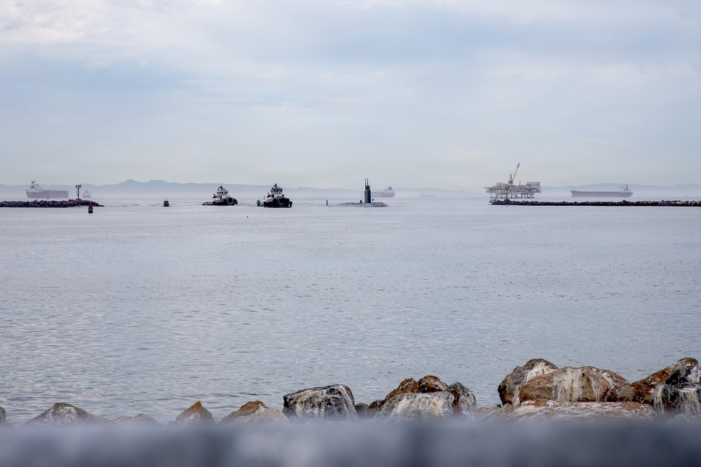 A harbor or bay scene photographed from a rocky breakwater showing multiple vessels on calm water under an overcast sky. The scene includes what appears to be a submarine being escorted by tugboats, along with an offshore oil platform being towed, and several large cargo or tanker ships in the background. Visibility is reduced by haze or marine fog on the horizon.