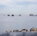 A harbor or bay scene photographed from a rocky breakwater showing multiple vessels on calm water under an overcast sky. The scene includes what appears to be a submarine being escorted by tugboats, along with an offshore oil platform being towed, and several large cargo or tanker ships in the background. Visibility is reduced by haze or marine fog on the horizon.