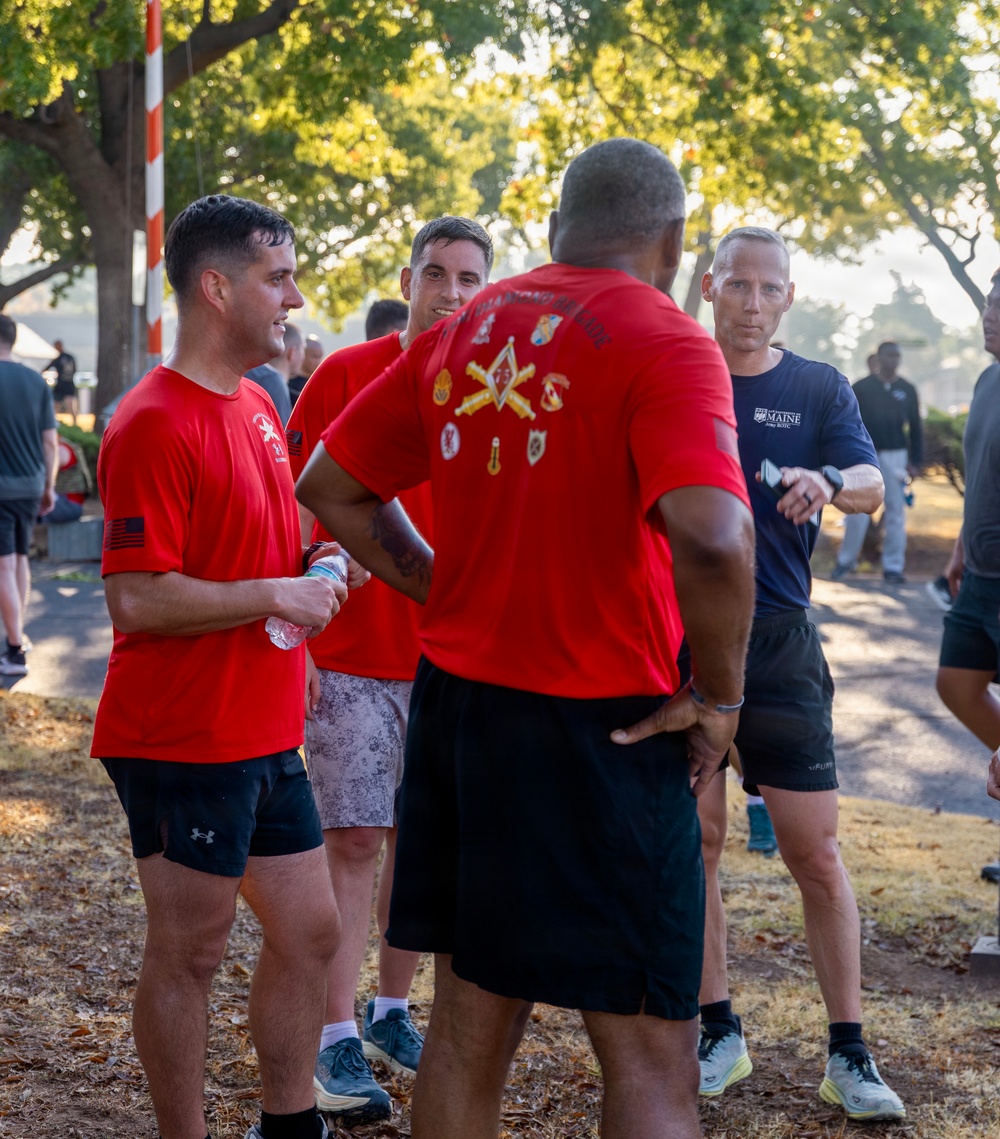 A group of individuals in athletic wear are gathered outdoors in what appears to be a military or ROTC physical training event. Several people are wearing matching red t-shirts with military insignia on the back, while one person in the background wears a University of Maine Army ROTC shirt. The setting appears to be a park or military installation with trees and a barber pole-style striped post visible in the background.