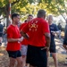 A group of individuals in athletic wear are gathered outdoors in what appears to be a military or ROTC physical training event. Several people are wearing matching red t-shirts with military insignia on the back, while one person in the background wears a University of Maine Army ROTC shirt. The setting appears to be a park or military installation with trees and a barber pole-style striped post visible in the background.
