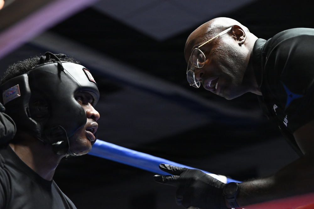 A boxing trainer leans over the ring ropes to speak with a boxer wearing a black protective headgear. The trainer, a bald man wearing glasses and a dark jacket, appears to be giving instructions or advice between rounds. The blue ring rope is prominently visible separating the two individuals.