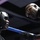 A boxing trainer leans over the ring ropes to speak with a boxer wearing a black protective headgear. The trainer, a bald man wearing glasses and a dark jacket, appears to be giving instructions or advice between rounds. The blue ring rope is prominently visible separating the two individuals.