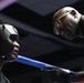 A boxing trainer leans over the ring ropes to speak with a boxer wearing a black protective headgear. The trainer, a bald man wearing glasses and a dark jacket, appears to be giving instructions or advice between rounds. The blue ring rope is prominently visible separating the two individuals.