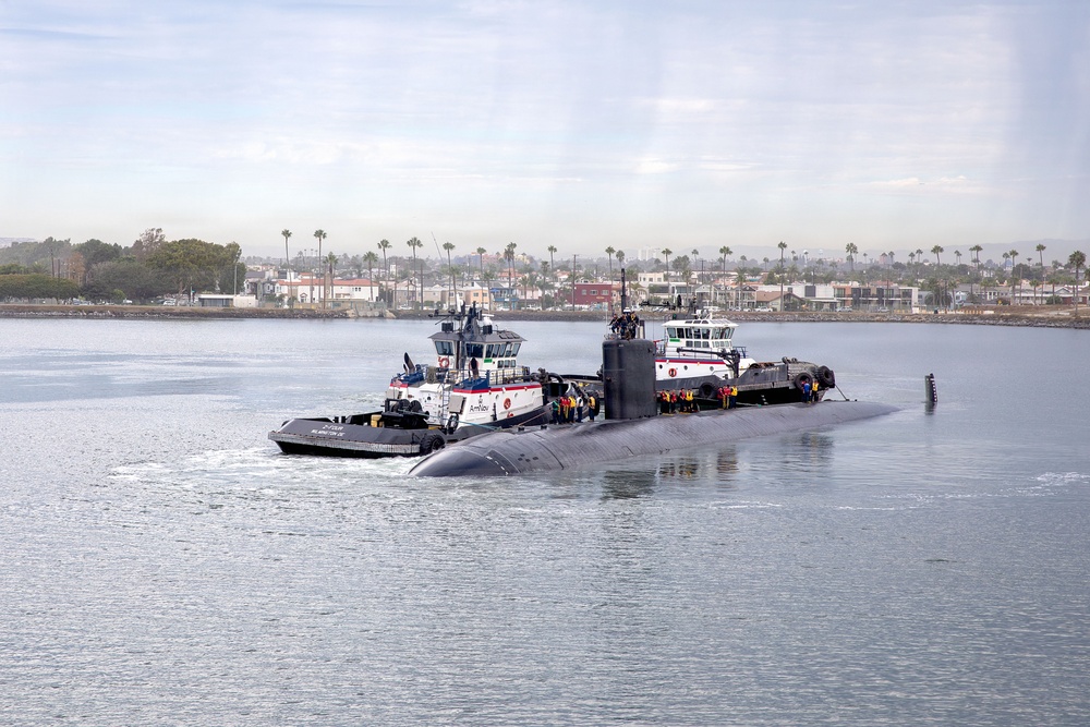A large dark-hulled military submarine is being assisted by two white tugboats in a harbor or bay. Personnel in colorful safety gear are visible standing on the submarine's deck. The background shows a coastal urban area with palm trees and low-rise buildings.