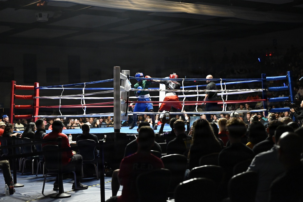 A live amateur boxing match is taking place in a well-lit ring surrounded by a large audience in an indoor venue. Two boxers are actively engaged in combat near a corner of the ring, with a referee closely monitoring the action. The crowd fills the surrounding area, watching the bout from ringside seats and bleachers in the background.