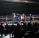 A live amateur boxing match is taking place in a well-lit ring surrounded by a large audience in an indoor venue. Two boxers are actively engaged in combat near a corner of the ring, with a referee closely monitoring the action. The crowd fills the surrounding area, watching the bout from ringside seats and bleachers in the background.