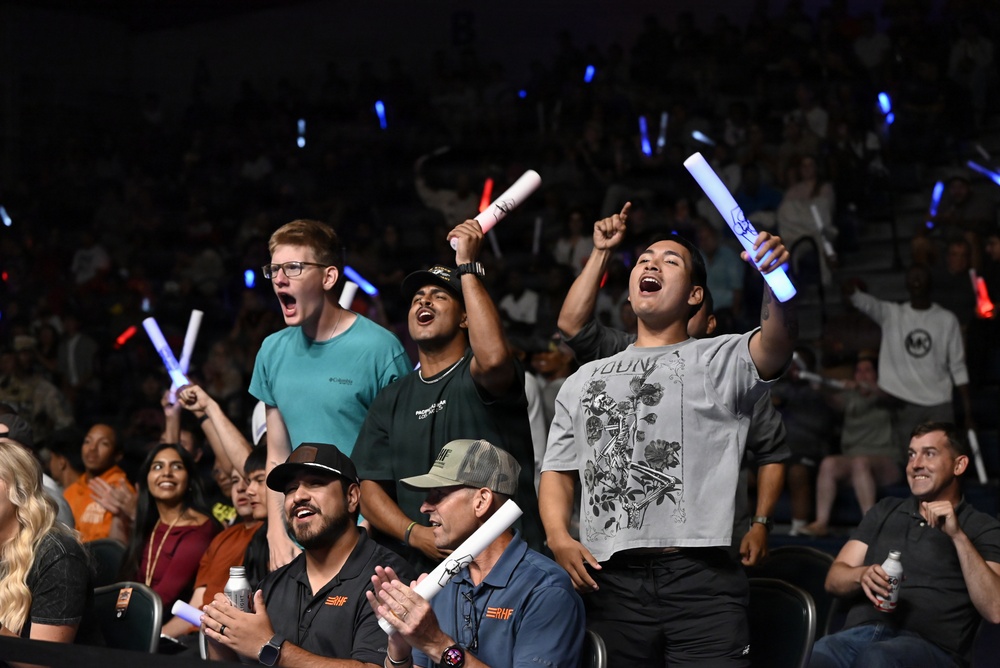 A crowd of enthusiastic spectators at an indoor arena event, cheering and celebrating with light-up foam sticks. Several people in the foreground are standing and shouting with excitement, while the background shows a packed audience with blue and red glowing sticks. Two individuals in the front row are wearing matching black shirts with an 'RHF' logo, suggesting possible staff or organizational affiliation.