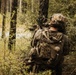 A soldier in full tactical gear is positioned in a wooded area, aiming a rifle toward an open clearing. The individual is viewed from behind and is partially concealed by dense vegetation. The scene appears to depict a military training exercise or field operation in a forest environment.