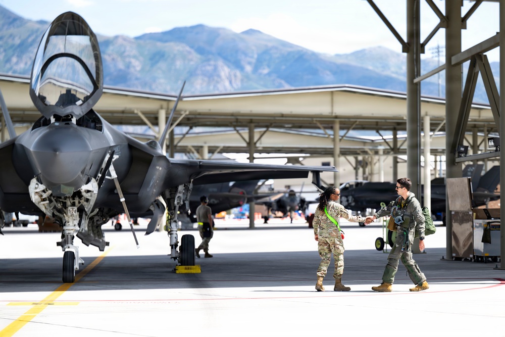 Two military personnel are shaking hands on an active flight line beside an F-35 Lightning II stealth fighter jet. The scene appears to take place at a military air base situated near a mountain range, likely in the western United States. Additional aircraft and ground crew are visible in the background under a large hangar structure.