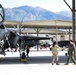 Two military personnel are shaking hands on an active flight line beside an F-35 Lightning II stealth fighter jet. The scene appears to take place at a military air base situated near a mountain range, likely in the western United States. Additional aircraft and ground crew are visible in the background under a large hangar structure.