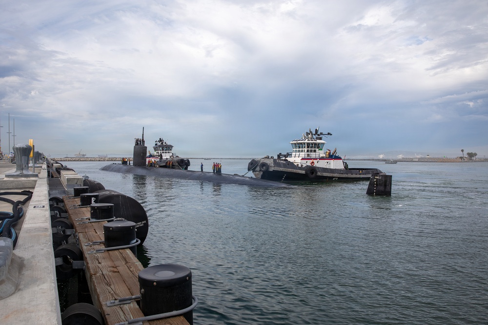A large dark-hulled military submarine is being guided into a naval pier with the assistance of at least one white tugboat positioned alongside it. Several crew members are visible standing on the submarine's deck during the docking operation. The scene takes place in a harbor with a concrete pier in the foreground and overcast skies above.