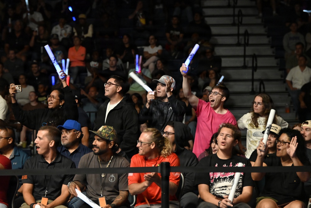 A crowd of enthusiastic spectators at an indoor arena event, with several fans in the front rows standing and cheering while holding illuminated foam glow sticks. The audience appears highly engaged, with visible expressions of excitement and joy. The setting appears to be a sporting or entertainment event held in a gymnasium or arena with bleacher seating.