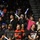 A crowd of enthusiastic spectators at an indoor arena event, with several fans in the front rows standing and cheering while holding illuminated foam glow sticks. The audience appears highly engaged, with visible expressions of excitement and joy. The setting appears to be a sporting or entertainment event held in a gymnasium or arena with bleacher seating.