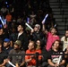 A crowd of enthusiastic spectators at an indoor arena event, with several fans in the front rows standing and cheering while holding illuminated foam glow sticks. The audience appears highly engaged, with visible expressions of excitement and joy. The setting appears to be a sporting or entertainment event held in a gymnasium or arena with bleacher seating.