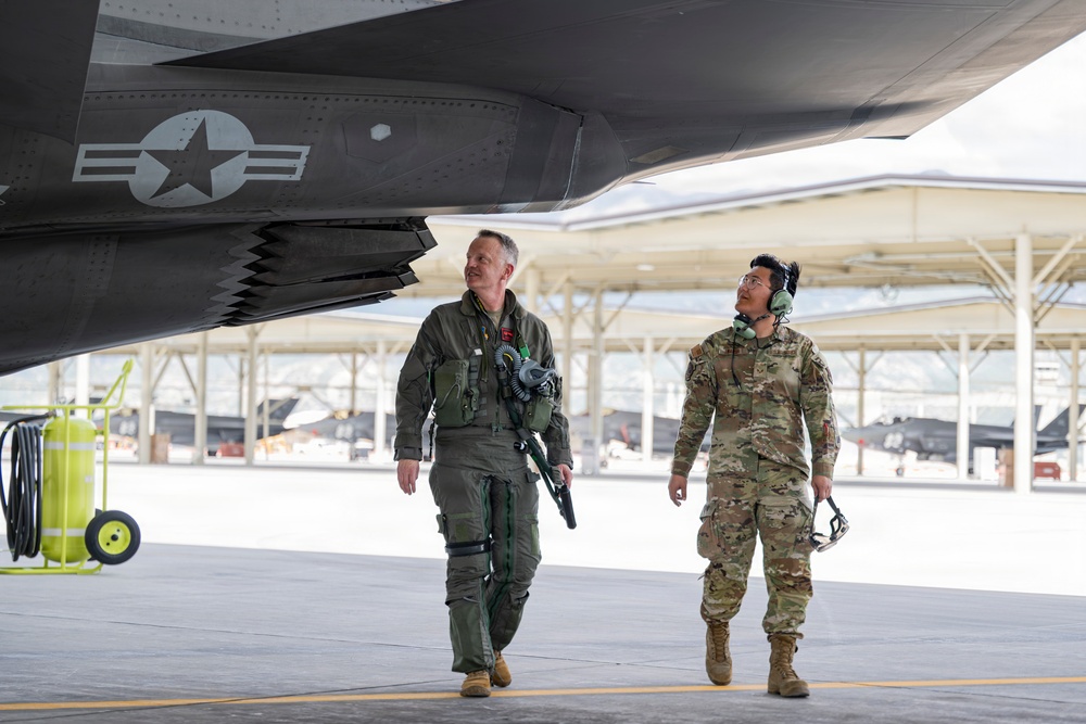 Two U.S. military personnel are walking beneath the wing of what appears to be an F-35 stealth fighter jet on a flight line ramp. One individual is wearing a pilot's flight suit with oxygen mask and G-suit attachments, while the other is in Army Combat Uniform (ACU/OCP) with hearing protection headset. Additional stealth aircraft are visible in the background near a hangar structure.