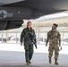 Two U.S. military personnel are walking beneath the wing of what appears to be an F-35 stealth fighter jet on a flight line ramp. One individual is wearing a pilot's flight suit with oxygen mask and G-suit attachments, while the other is in Army Combat Uniform (ACU/OCP) with hearing protection headset. Additional stealth aircraft are visible in the background near a hangar structure.