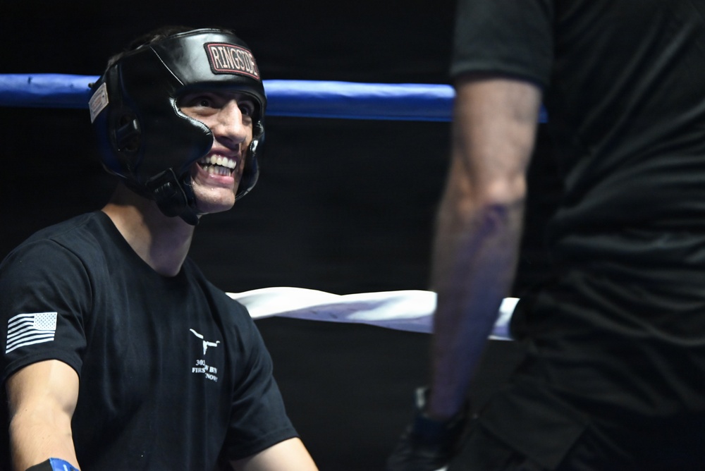 A smiling young person wearing a black Ringside brand boxing headgear is positioned inside a boxing ring, appearing relaxed and cheerful. They are wearing a black t-shirt with an American flag patch on the sleeve and what appears to be a longhorn graphic on the chest. A second partially visible person stands to the right, and blue ring ropes are visible in the background.