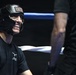 A smiling young person wearing a black Ringside brand boxing headgear is positioned inside a boxing ring, appearing relaxed and cheerful. They are wearing a black t-shirt with an American flag patch on the sleeve and what appears to be a longhorn graphic on the chest. A second partially visible person stands to the right, and blue ring ropes are visible in the background.