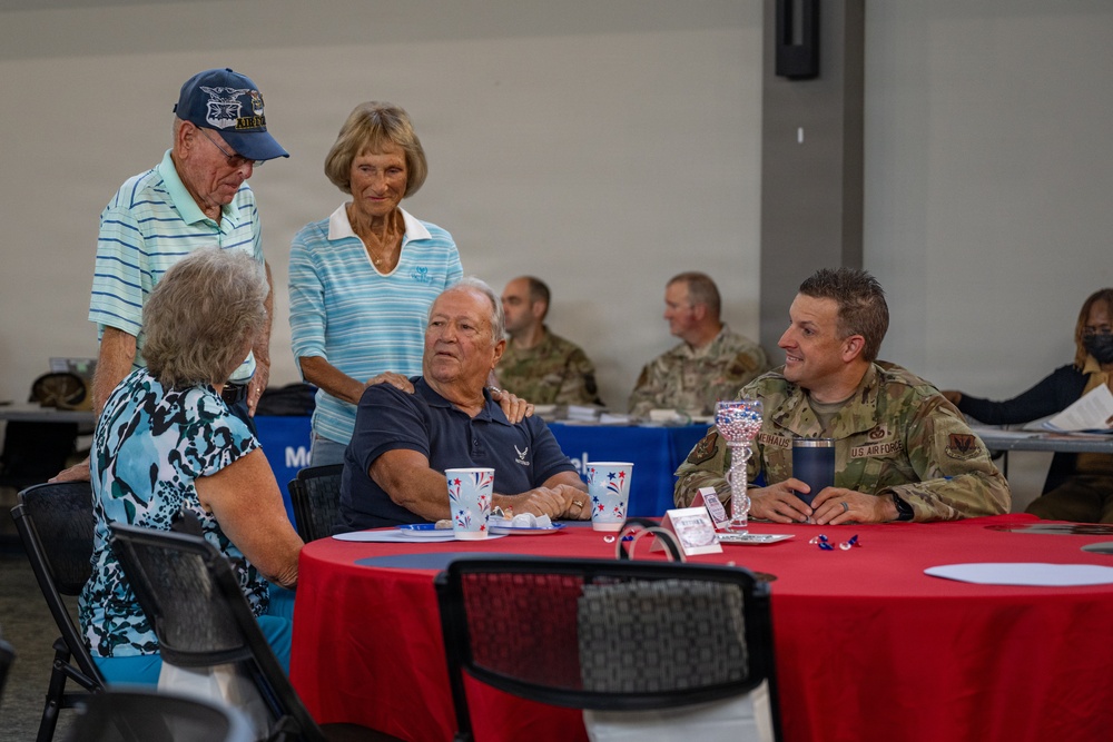 A group of civilians and U.S. Air Force military personnel are gathered at what appears to be a veterans or military appreciation event, seated and standing around a red-clothed round table. The setting includes patriotic decorations such as red, white, and blue cups and table accessories. Interactions appear warm and social, suggesting a community outreach or recognition ceremony.