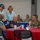 A group of civilians and U.S. Air Force military personnel are gathered at what appears to be a veterans or military appreciation event, seated and standing around a red-clothed round table. The setting includes patriotic decorations such as red, white, and blue cups and table accessories. Interactions appear warm and social, suggesting a community outreach or recognition ceremony.