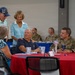 A group of civilians and U.S. Air Force military personnel are gathered at what appears to be a veterans or military appreciation event, seated and standing around a red-clothed round table. The setting includes patriotic decorations such as red, white, and blue cups and table accessories. Interactions appear warm and social, suggesting a community outreach or recognition ceremony.