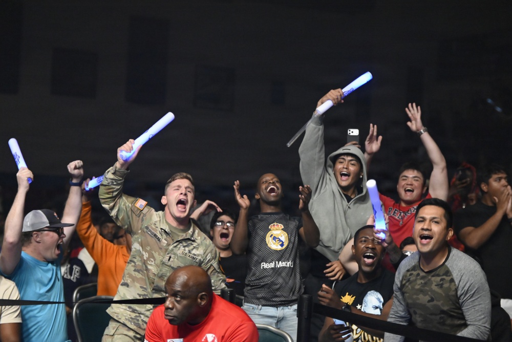 A group of enthusiastic fans are cheering at what appears to be a nighttime sporting event. Several individuals are holding glowing blue foam sticks and raising their arms in celebration. Notable attendees include a U.S. Army soldier in camouflage uniform and a person wearing a Real Madrid jersey.