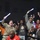 A group of enthusiastic fans are cheering at what appears to be a nighttime sporting event. Several individuals are holding glowing blue foam sticks and raising their arms in celebration. Notable attendees include a U.S. Army soldier in camouflage uniform and a person wearing a Real Madrid jersey.