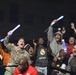 A group of enthusiastic fans are cheering at what appears to be a nighttime sporting event. Several individuals are holding glowing blue foam sticks and raising their arms in celebration. Notable attendees include a U.S. Army soldier in camouflage uniform and a person wearing a Real Madrid jersey.