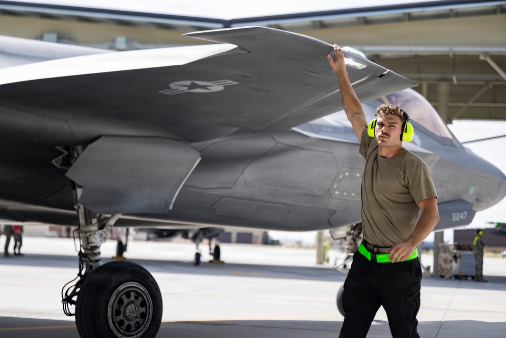 A military aircraft maintainer or crew chief is conducting a pre-flight or post-flight inspection of what appears to be an F-35 Lightning II stealth fighter jet on a flight line. The technician is wearing hearing protection and a high-visibility belt, touching the aircraft's wing surface while surveying the area. The setting appears to be a military air base with additional aircraft and personnel visible in the background.
