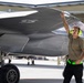 A military aircraft maintainer or crew chief is conducting a pre-flight or post-flight inspection of what appears to be an F-35 Lightning II stealth fighter jet on a flight line. The technician is wearing hearing protection and a high-visibility belt, touching the aircraft's wing surface while surveying the area. The setting appears to be a military air base with additional aircraft and personnel visible in the background.